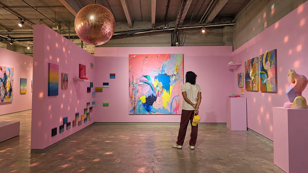 Man in burgundy trousers viewing abstract art in a pink gallery with a rose-gold disco ball and pastel sculpture.