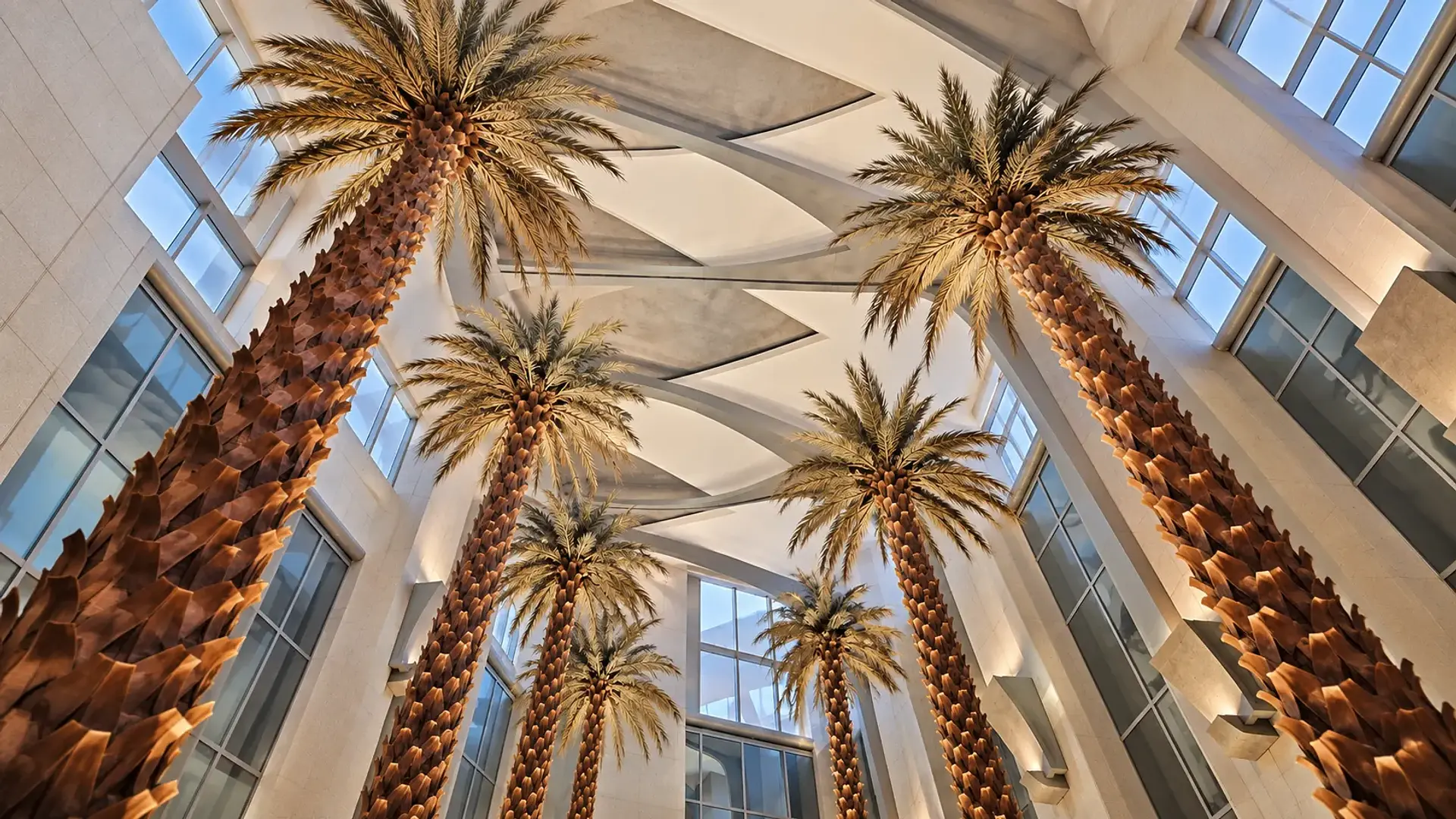 Upward view of tall date palms beneath a sculptural atrium ceiling, framed by pale stone walls and windows.